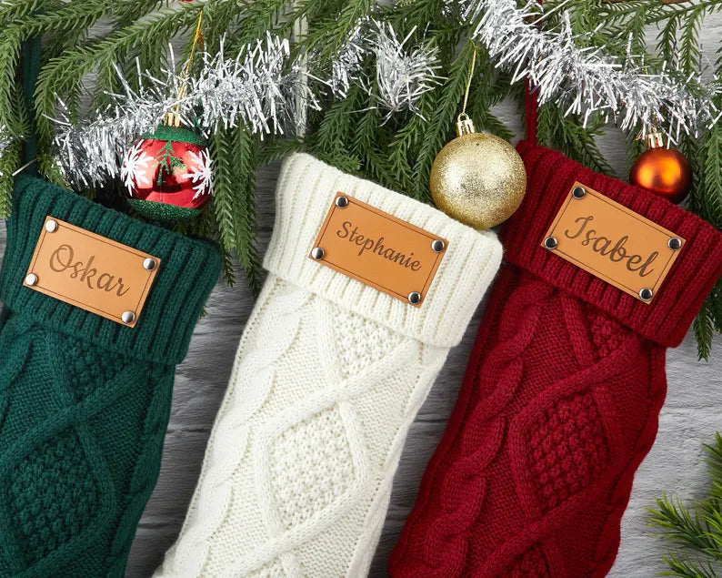 Set of personalized engraved leather Christmas stockings in green, white, and red, hanging on a festive tree branch with ornaments