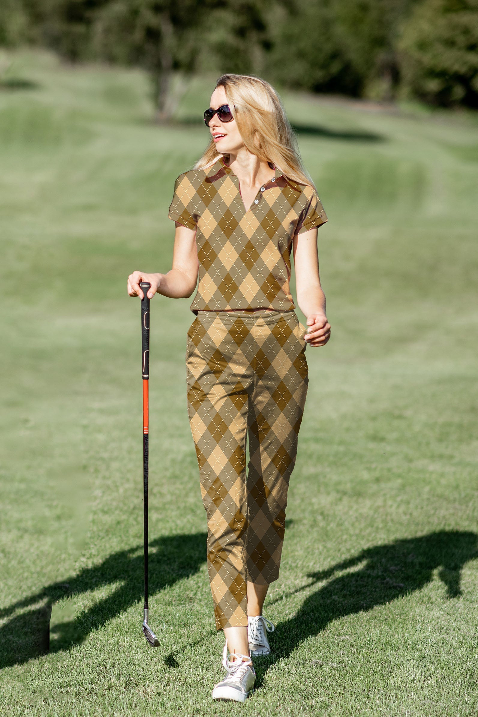 Woman wearing brown argyle women's golf polo and pants set on golf course with sunglasses and golf club